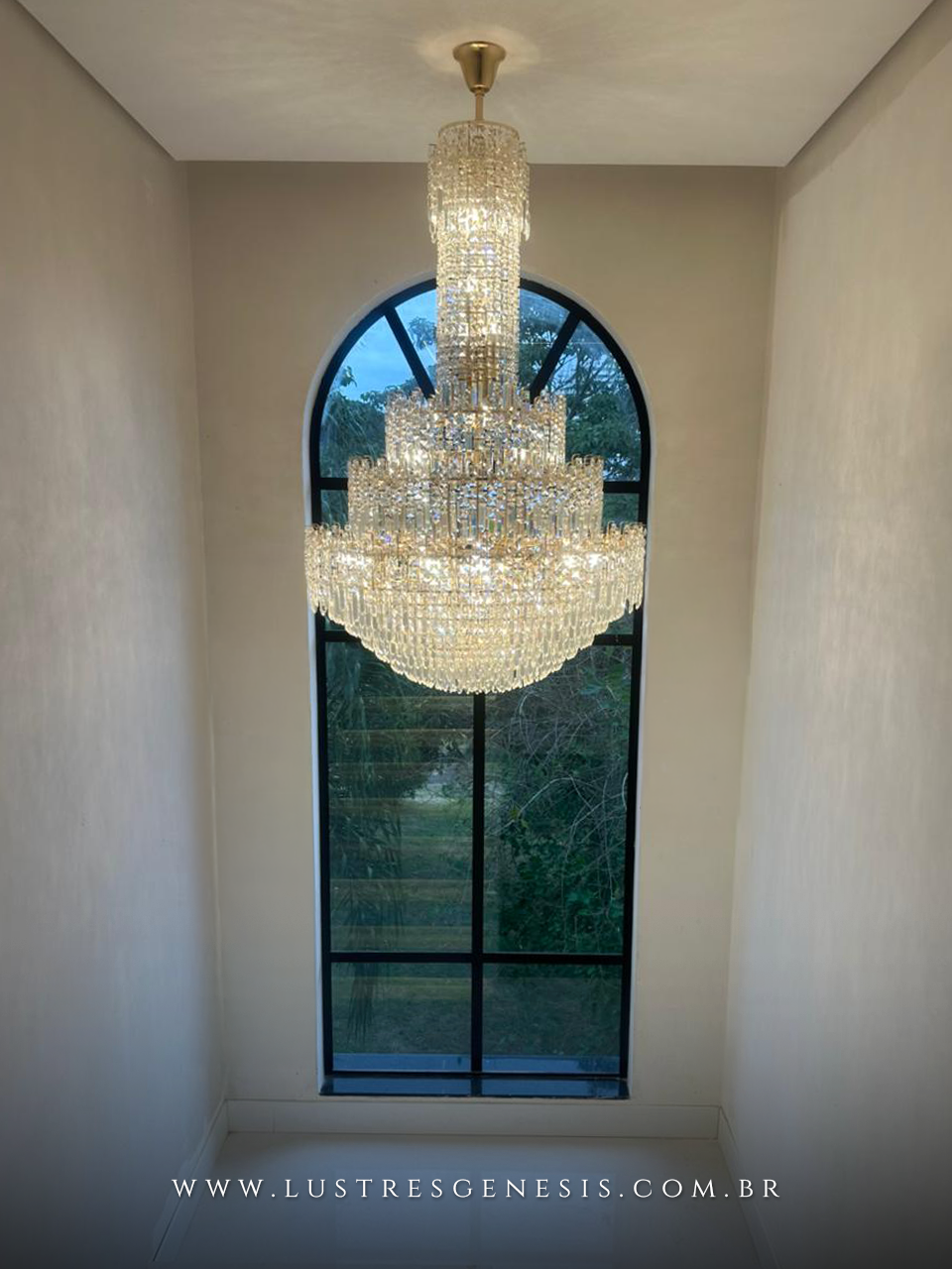 Lustre pendente de cristal grande modelo Meca dourado, instalado em hall com pé direito duplo e janela alta ao fundo.